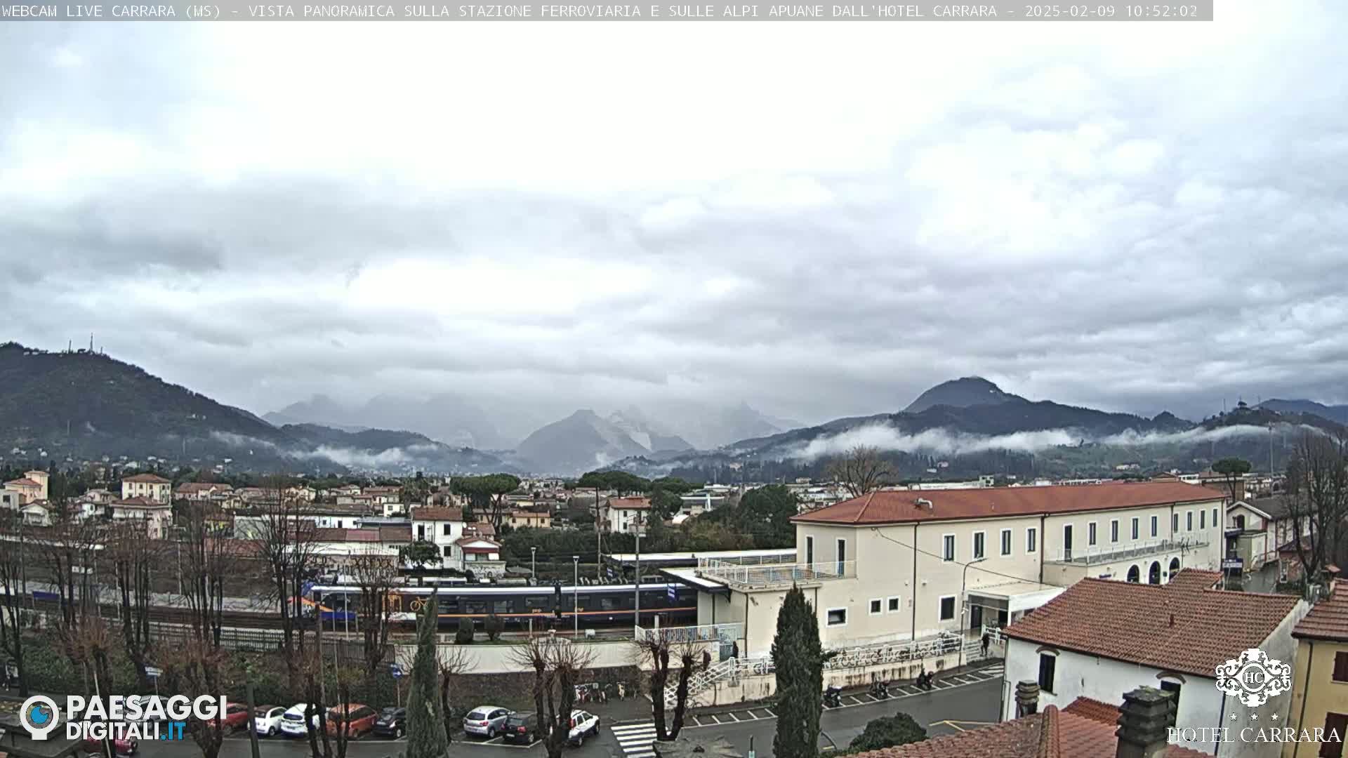 A train sits at a station in a town nestled in a valley, with misty mountains in the background under an overcast sky.
