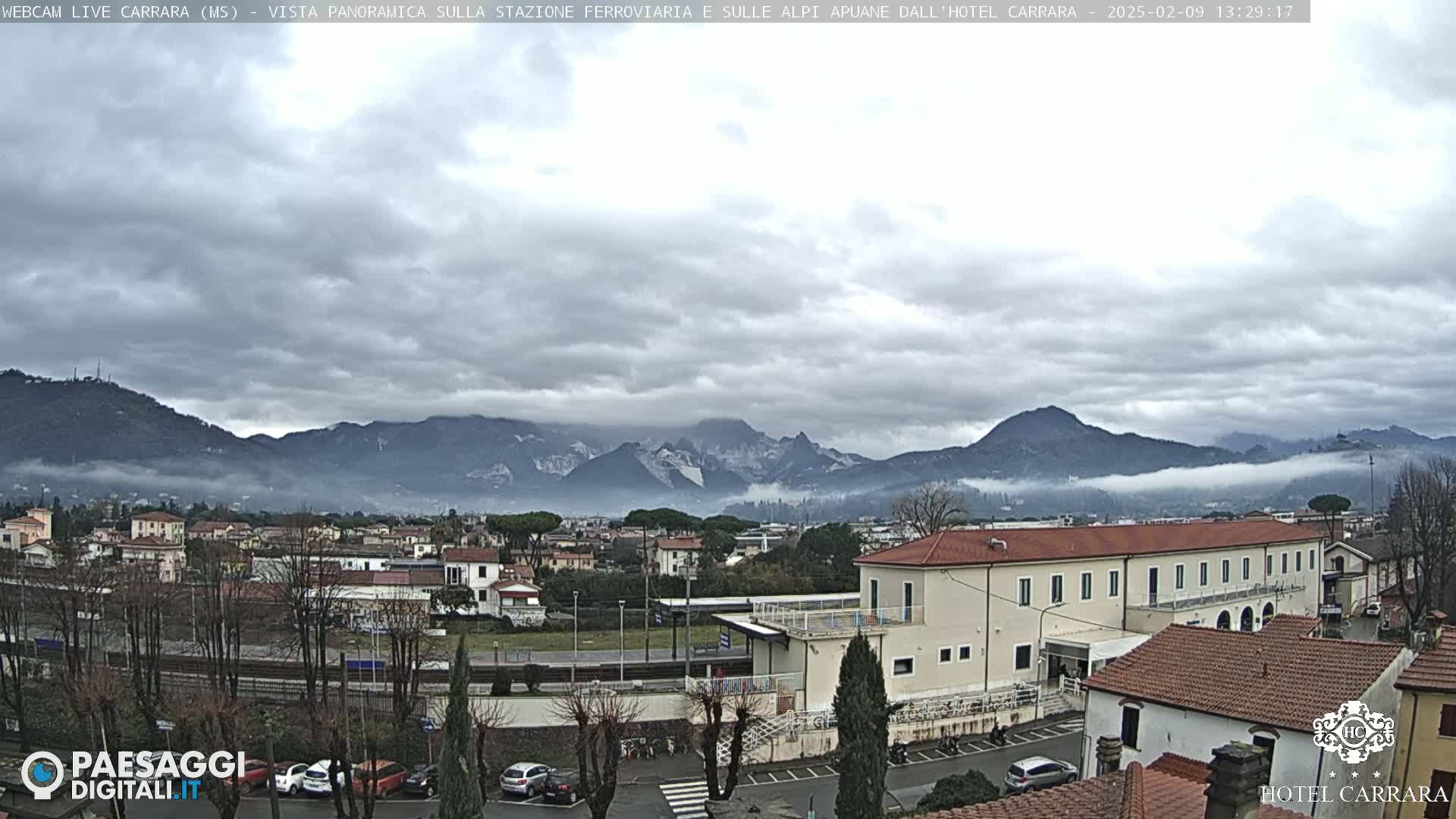 A cloudy day reveals a town nestled in a valley, with a train station and buildings in the foreground, and snow-capped mountains shrouded in fog in the background.