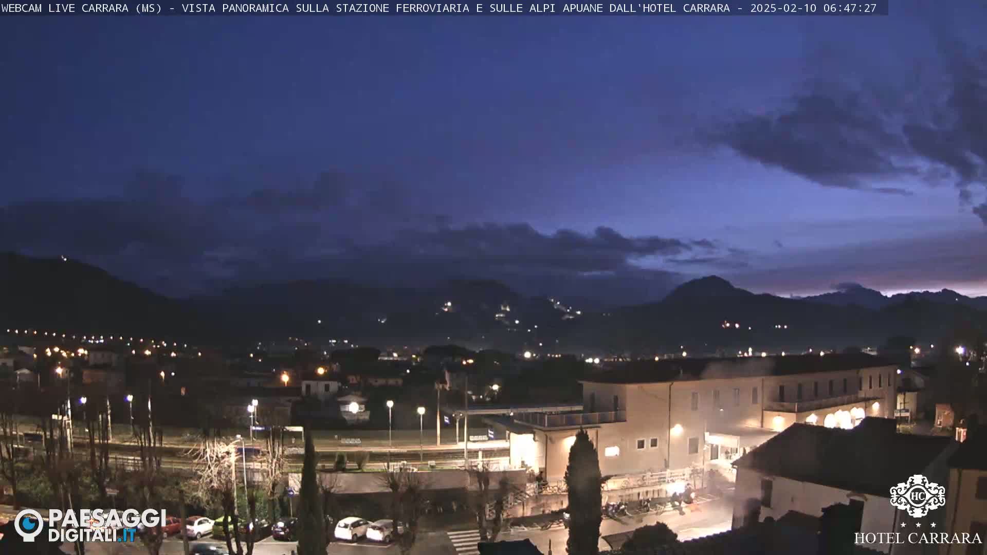 A twilight view of a town nestled in a valley, with mountains in the background under a mostly clear, dark blue sky.