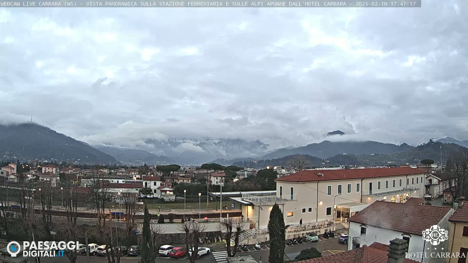 A cloudy, overcast day reveals a view of a town nestled beside mountains, with a train station and a hotel prominently featured in the foreground.