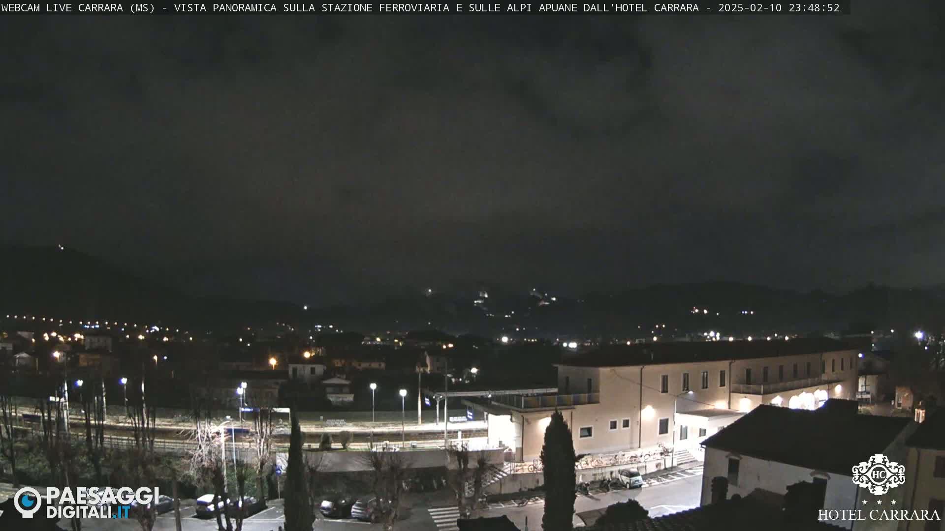 A nighttime panoramic view of a town, illuminated by streetlights, with a large building in the foreground and hills in the background under a cloudy sky.