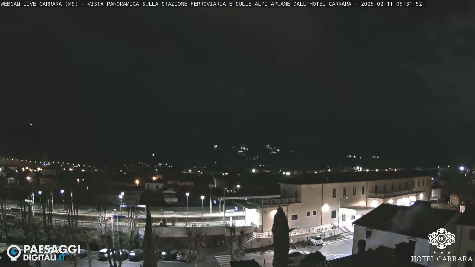 A nighttime long shot of a town's outskirts, featuring a train station and a hotel, under a dark, clear sky.