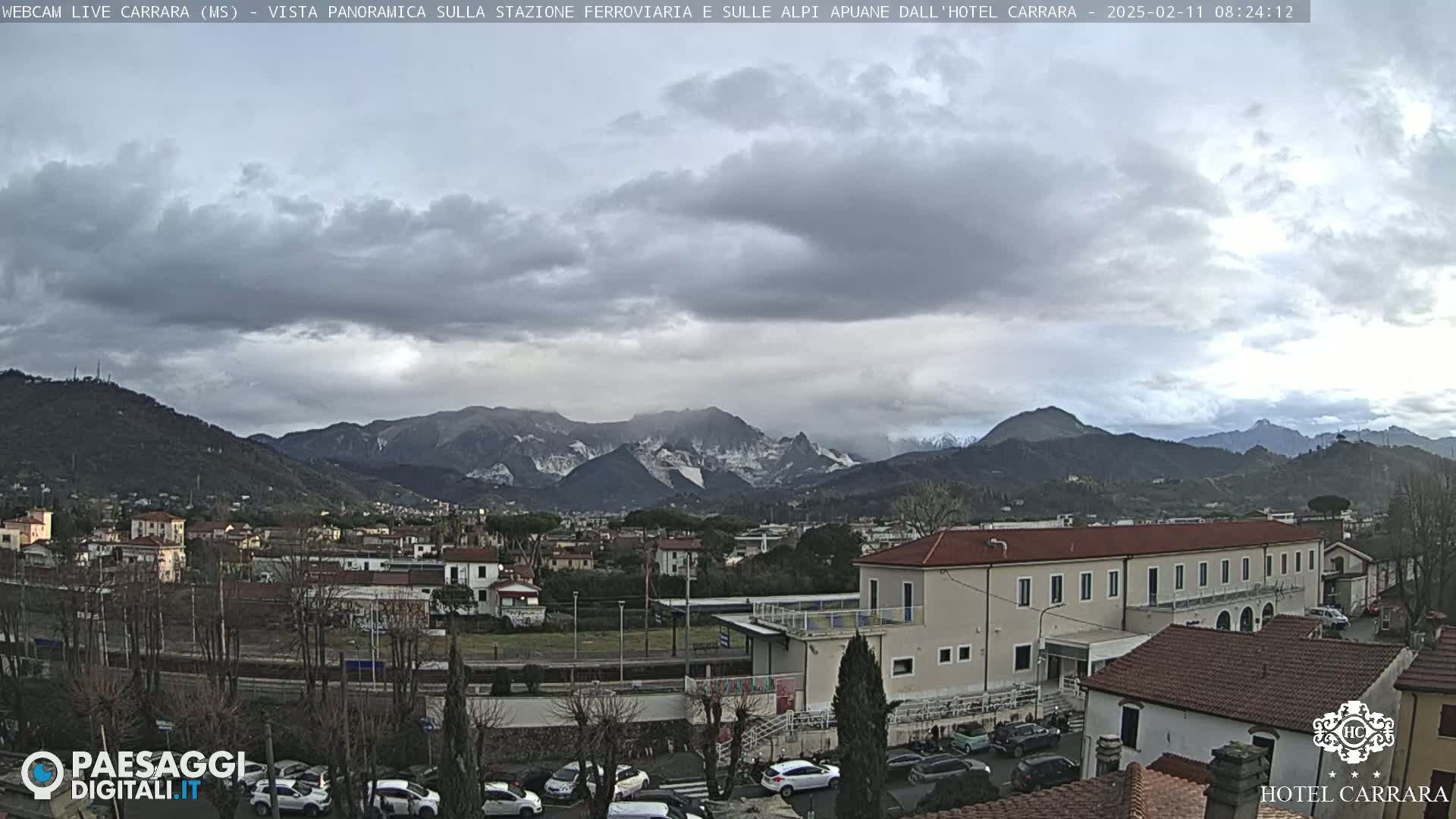 A panoramic view of a town nestled in a valley, with overcast skies and snow-capped mountains in the background.