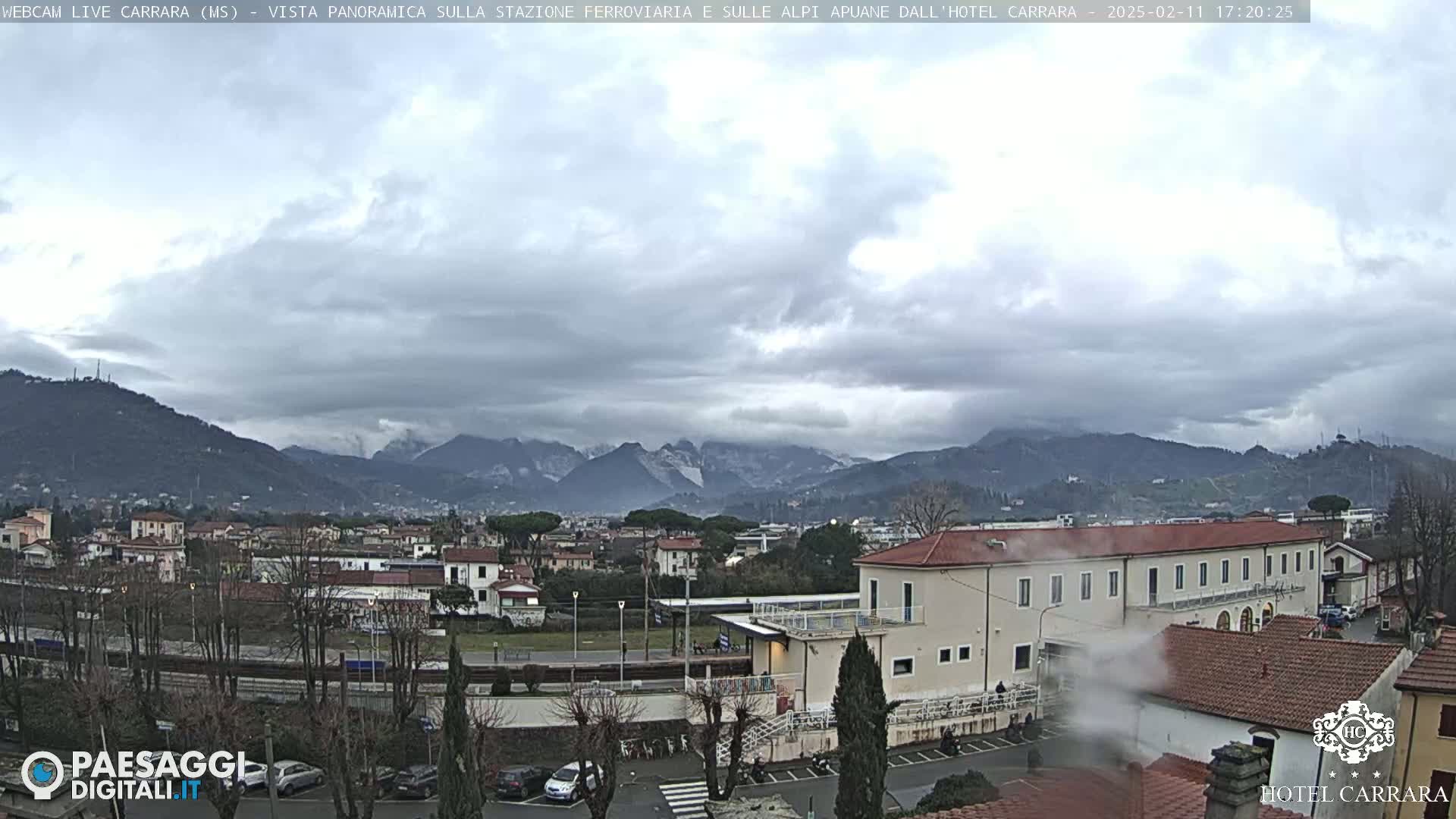 A cloudy, overcast day reveals a panoramic view of a town nestled between hills and mountains, with a train station and hotel visible in the foreground.