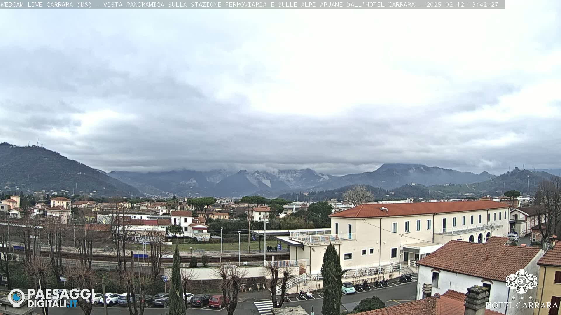 A cloudy day reveals a panoramic view of a town nestled beside mountains, with a train station and buildings visible in the foreground.