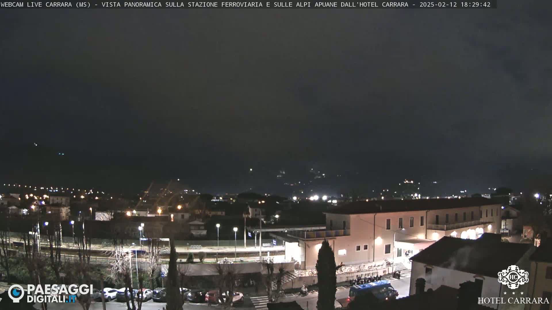 A nighttime panoramic view of a town with a train station and hotel, under a dark, cloudy sky.