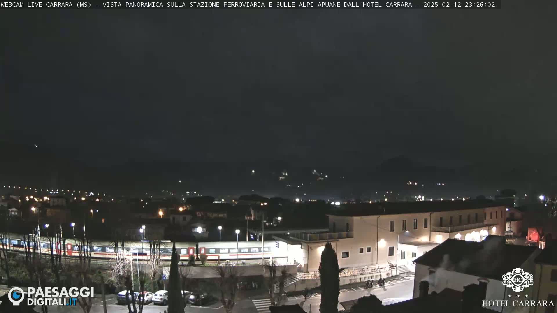 A nighttime, panoramic view shows a train at a station near a hotel and town, with dark mountains in the distance under a cloudy sky.