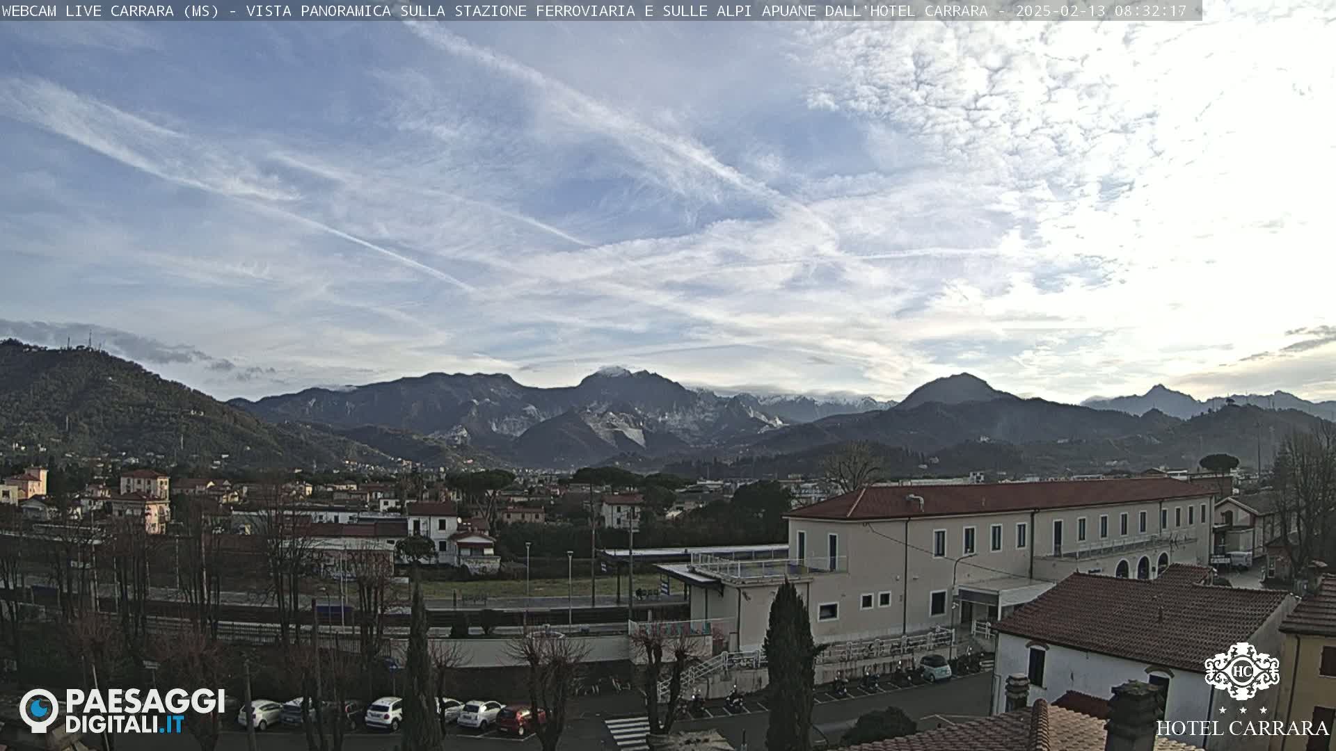 A panoramic view of a town nestled in a valley, with snow-capped mountains in the background under a mostly clear sky with some scattered clouds and contrails.