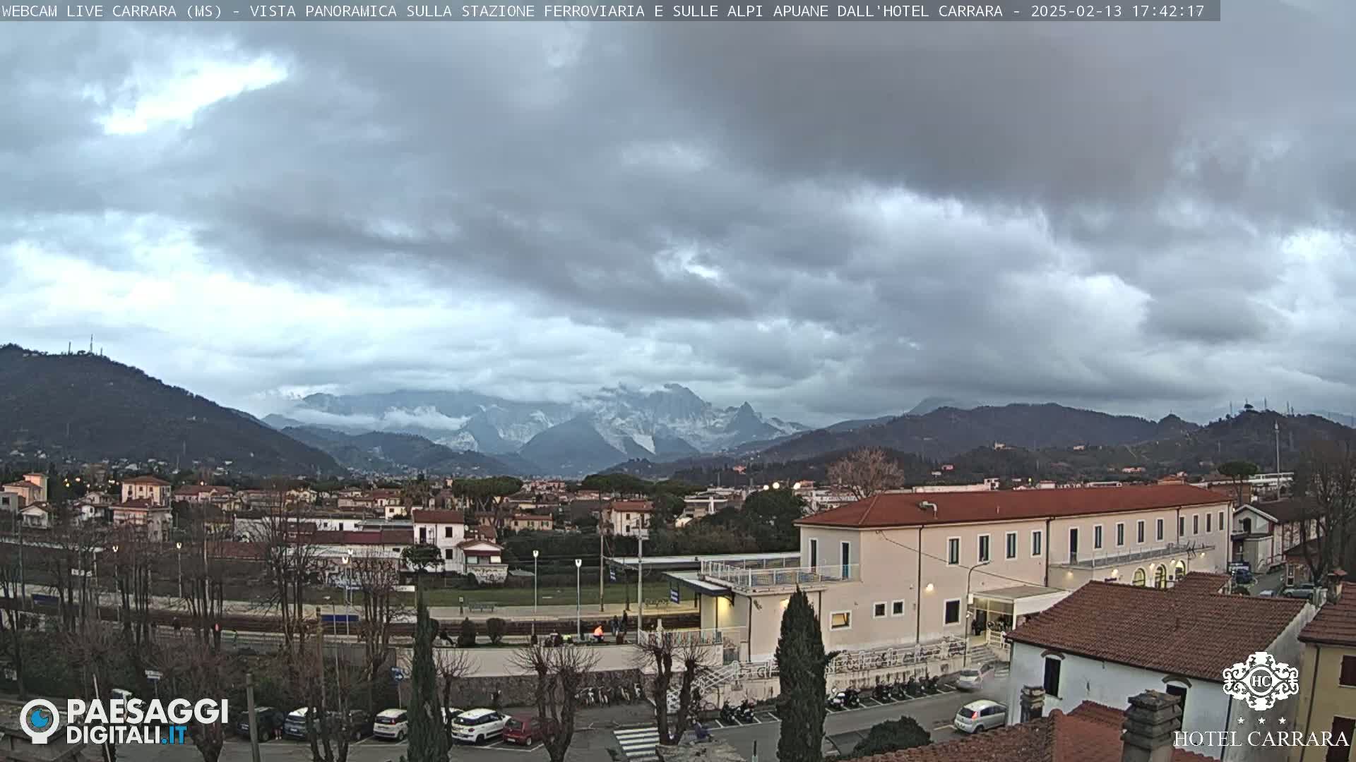 A panoramic view of a town nestled in a valley, with overcast skies and snow-capped mountains in the distance.