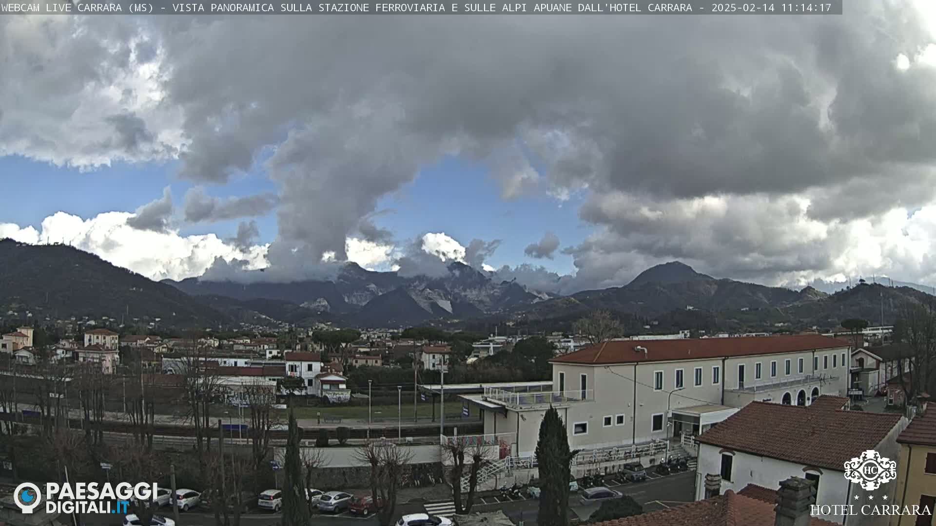 A partly cloudy day reveals a view of a town nestled among mountains, with a building and parking lot in the foreground.