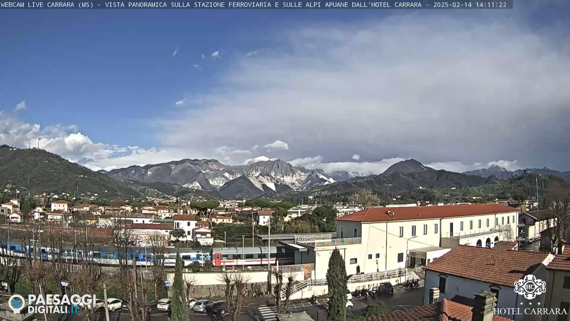 A partly cloudy day reveals a train station and town nestled in a valley, with snow-capped mountains in the background.