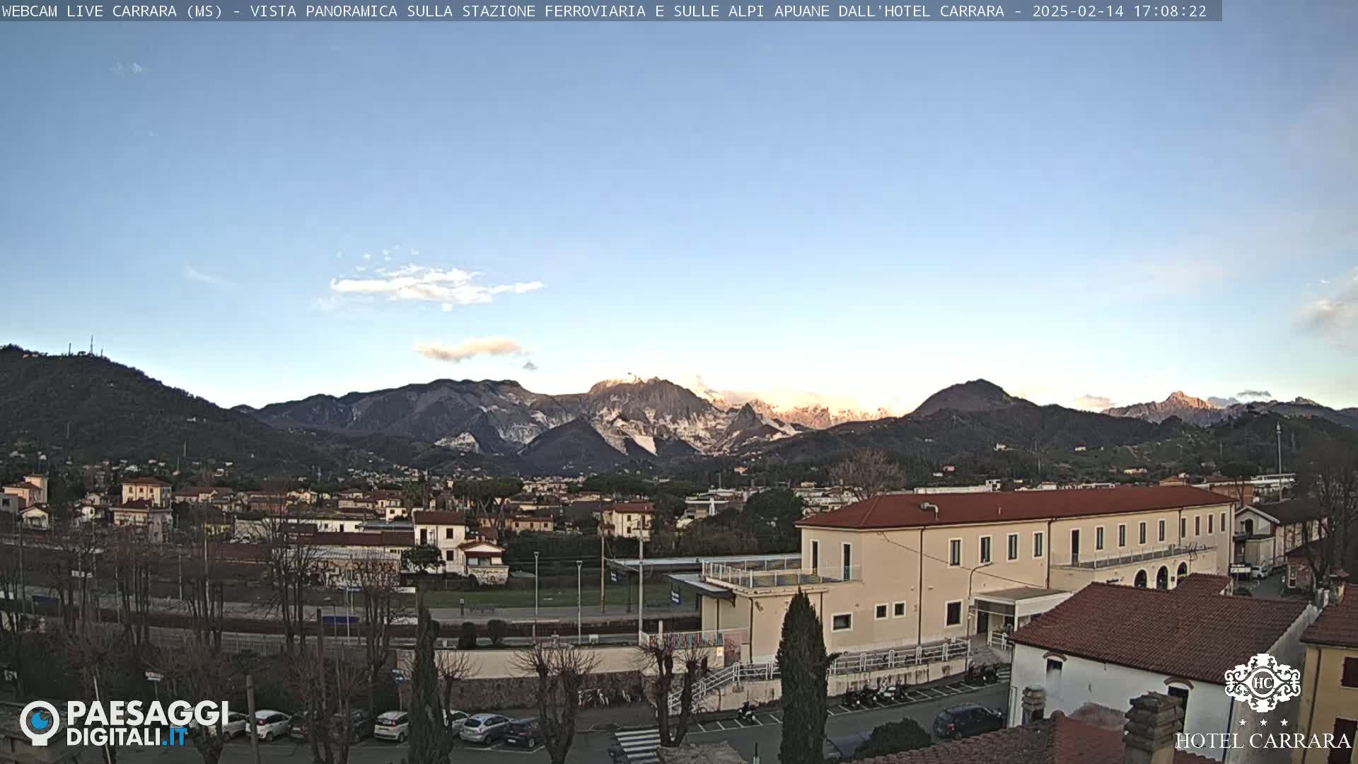 A long, low building sits in a town nestled at the foot of snow-capped mountains under a mostly clear, pale blue sky.