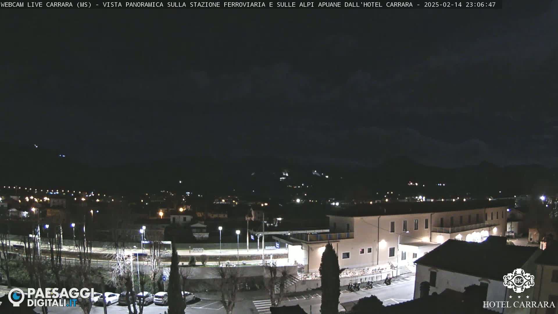 A nighttime, panoramic view of a town with a train station and a large building, illuminated by streetlights, with dark mountains in the background under a partly cloudy sky.