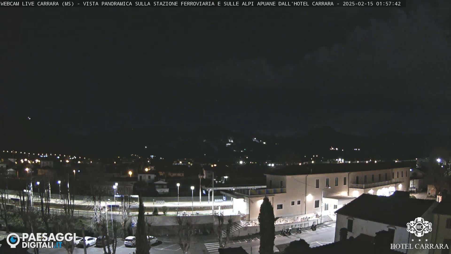A nighttime, slightly cloudy view of a town with a hotel and train station, nestled at the base of dark mountains.
