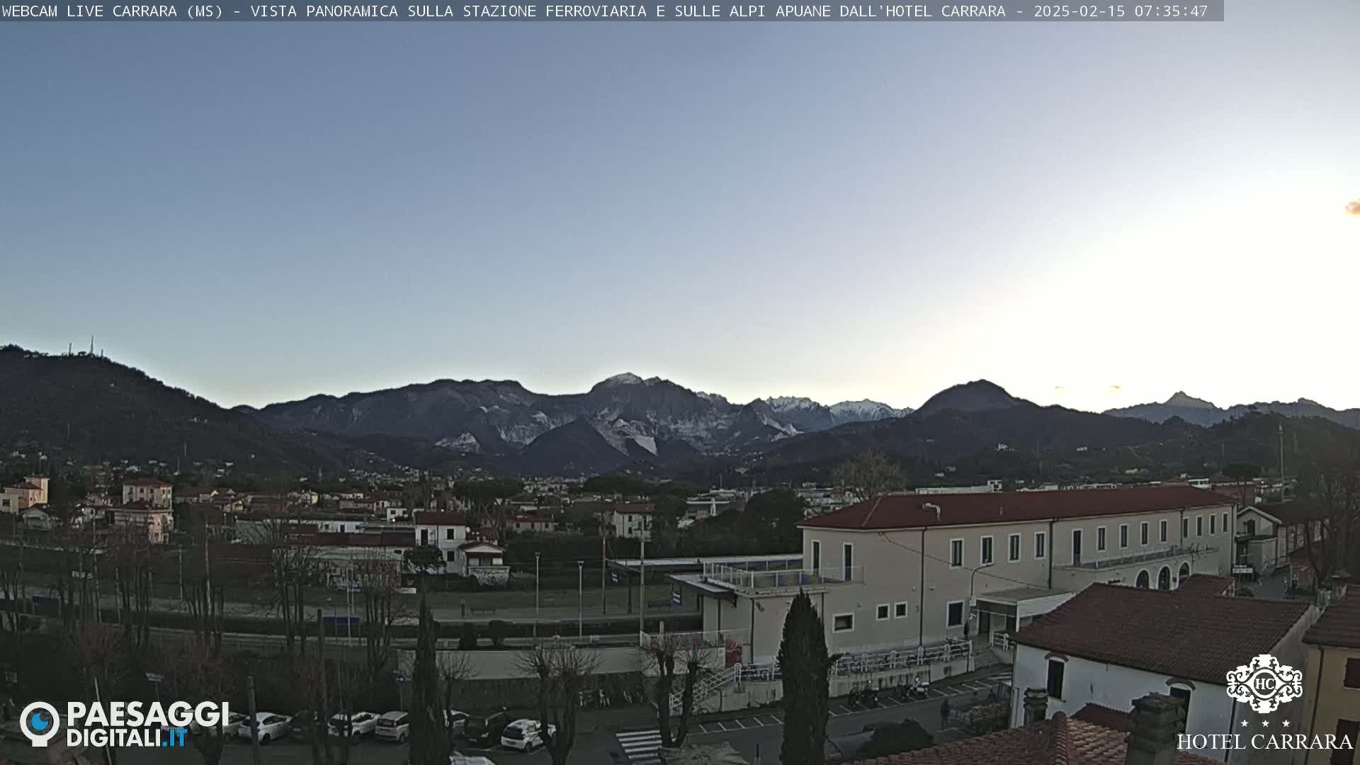 A panoramic view of a town nestled in a valley, with snow-capped mountains in the background under a clear, twilight sky.