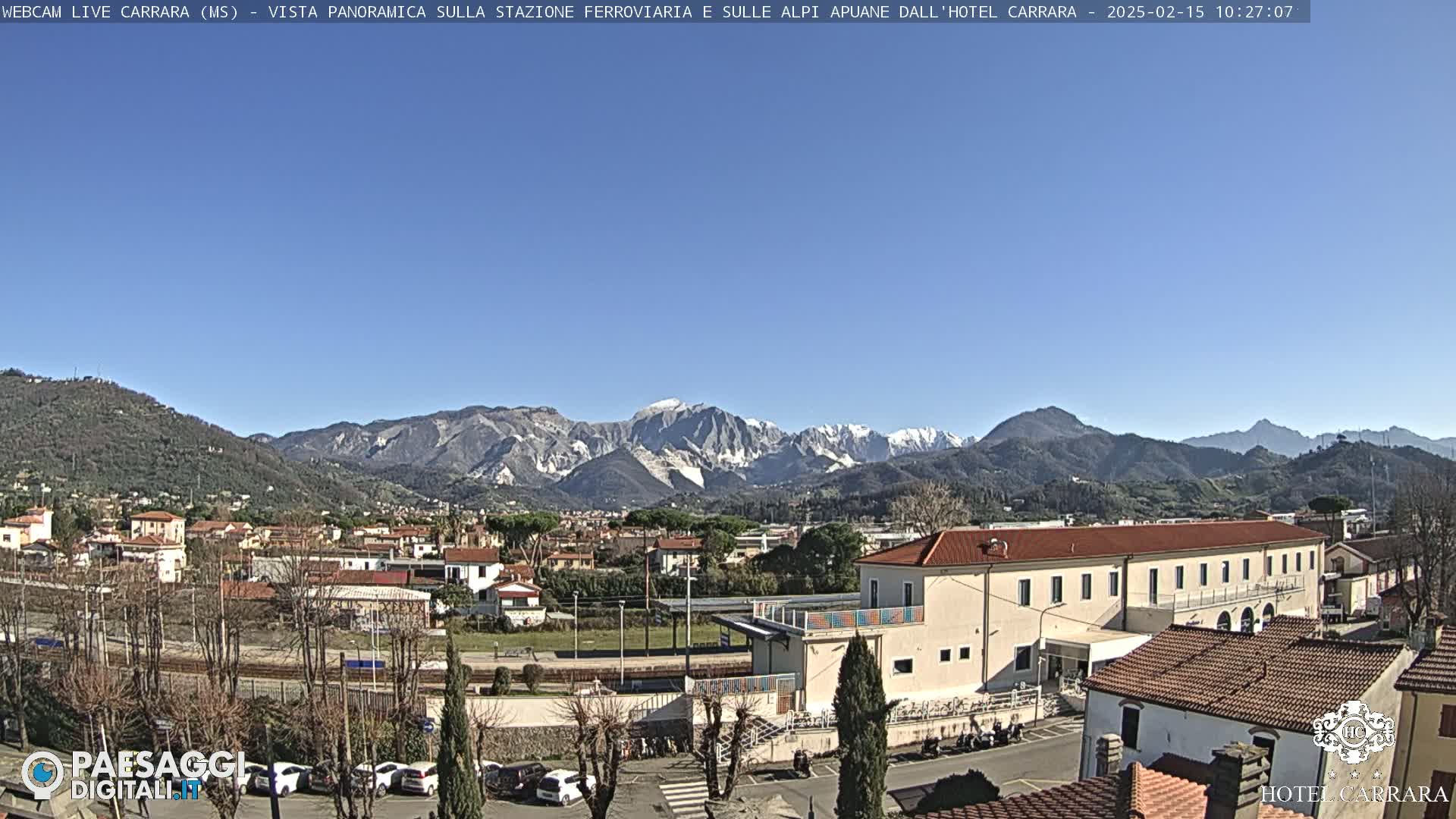 A clear, sunny day reveals a town nestled among rolling hills, with snow-capped mountains in the background.