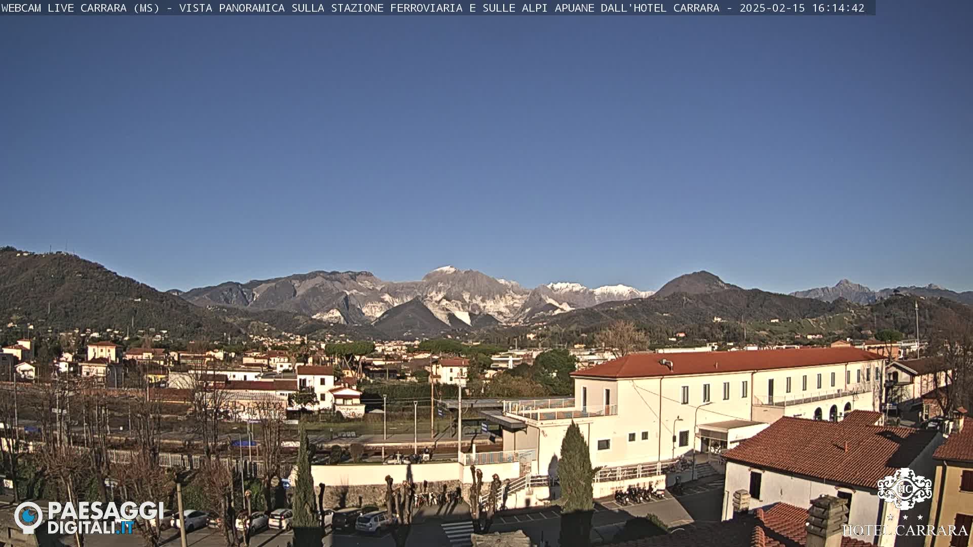 A long, low building with a red roof sits in a town nestled in a valley, with snow-capped mountains visible in the background under a clear blue sky.