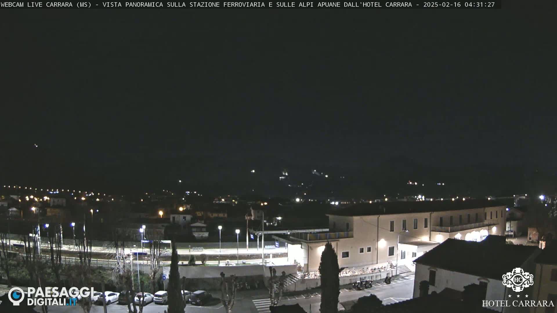 A nighttime long shot of a town's outskirts, featuring a train station and a hotel, under a clear, dark sky.