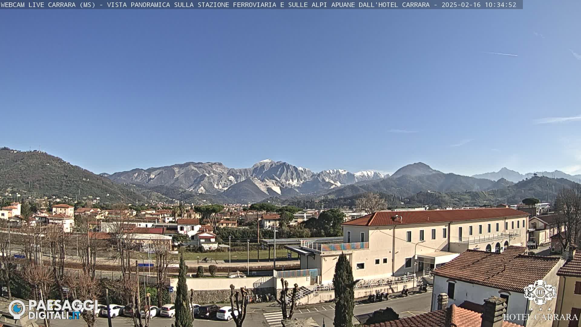 A clear, sunny day reveals a town nestled at the foot of snow-capped mountains, with a train station and buildings visible in the foreground.