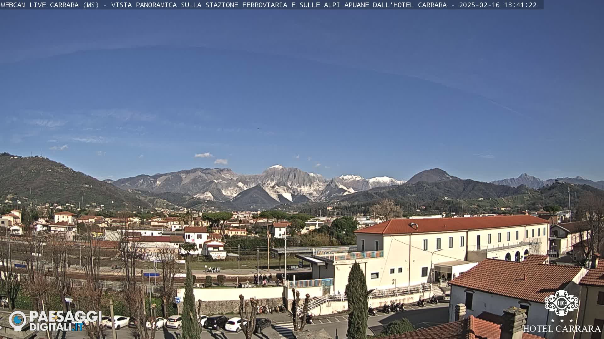A sunny day reveals a town nestled in a valley, with snow-capped mountains in the background.