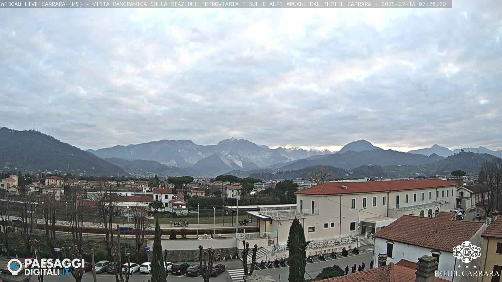 A cloudy day reveals a view of a town nestled in a valley, with snow-capped mountains in the background.