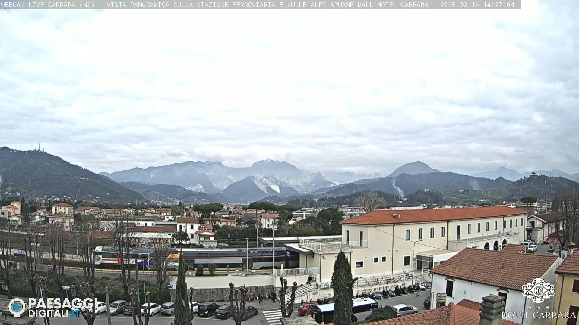 A train sits at a station in a town nestled in a valley, with mountains in the background under a cloudy sky.