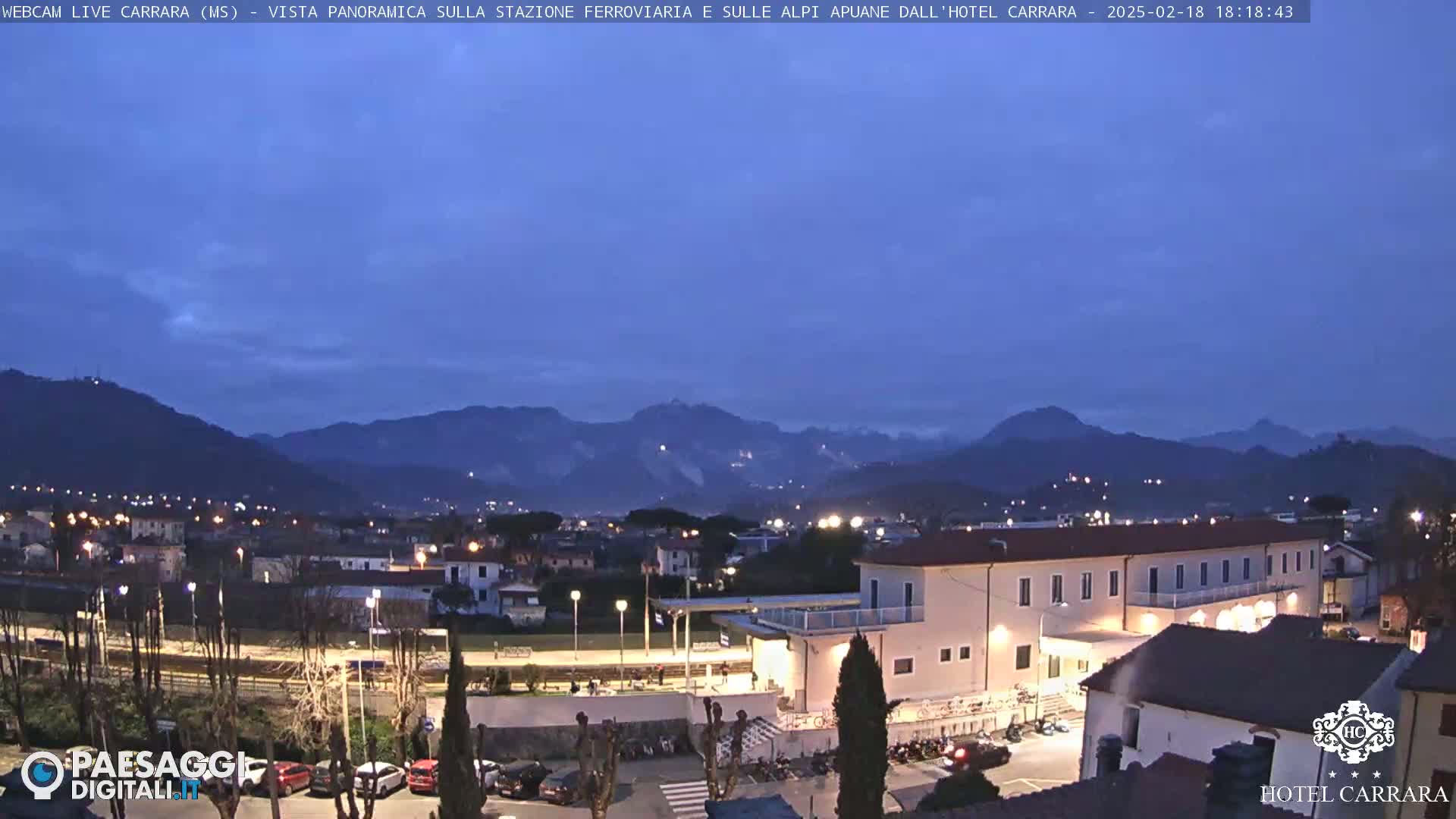 A twilight view of a town nestled in a valley, with mountains in the background under a mostly clear, dark blue sky.