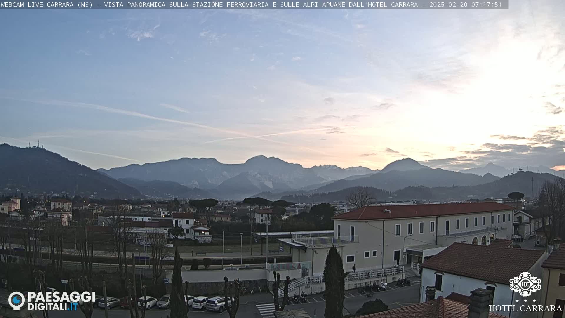 A panoramic view of a town nestled in a valley, overlooked by a mountain range under a pale sunrise sky.