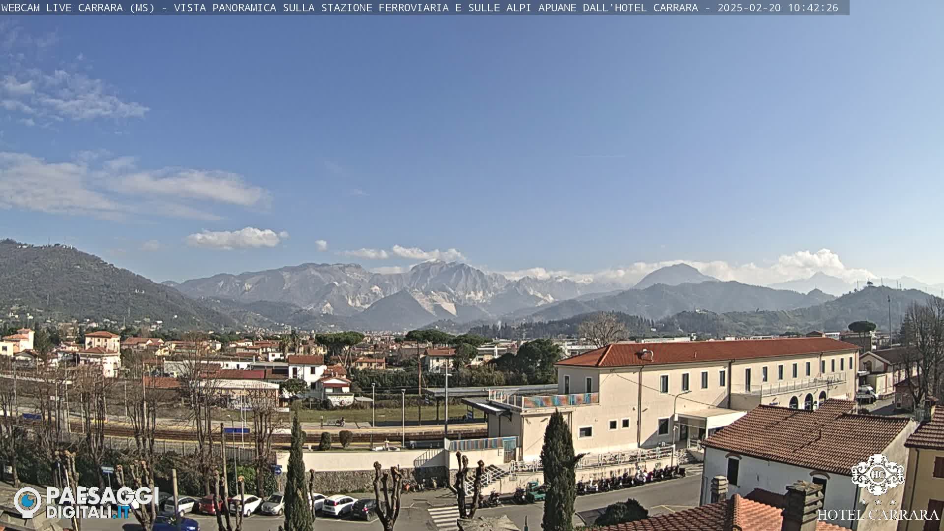 A sunny day reveals a panoramic view of a town nestled at the foot of snow-capped mountains, with a train station and buildings in the foreground.