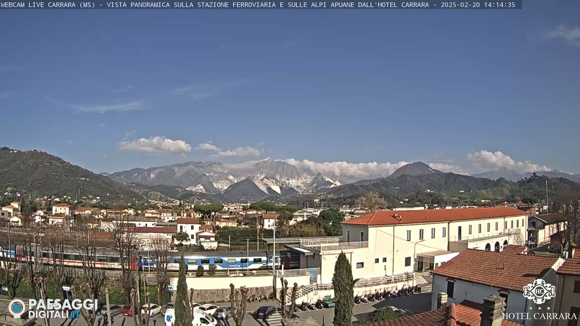 A long passenger train is visible near a large building, all situated in a town nestled at the foot of a mountain range under a mostly clear blue sky with a few small clouds.