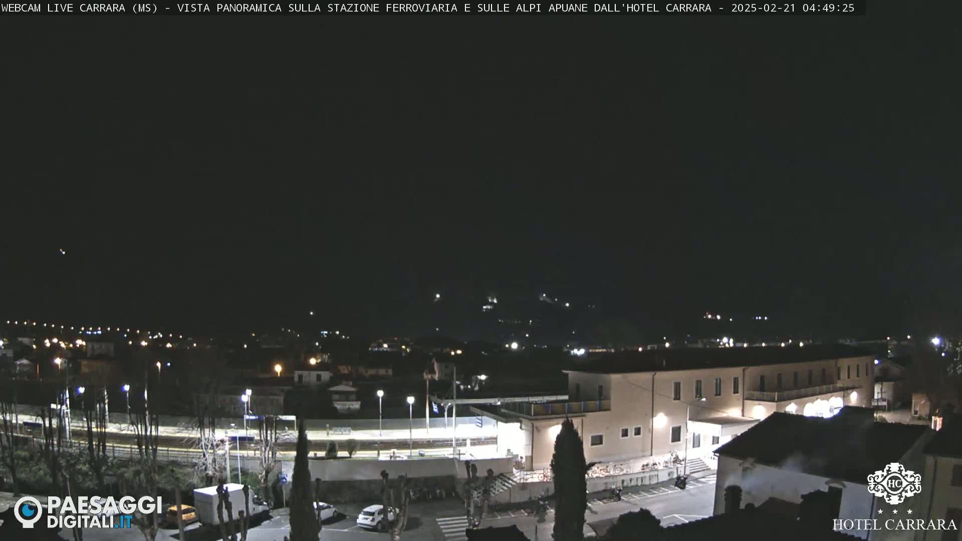 A nighttime panoramic view of a town, showing a train station, buildings, and distant hills under a clear, dark sky.