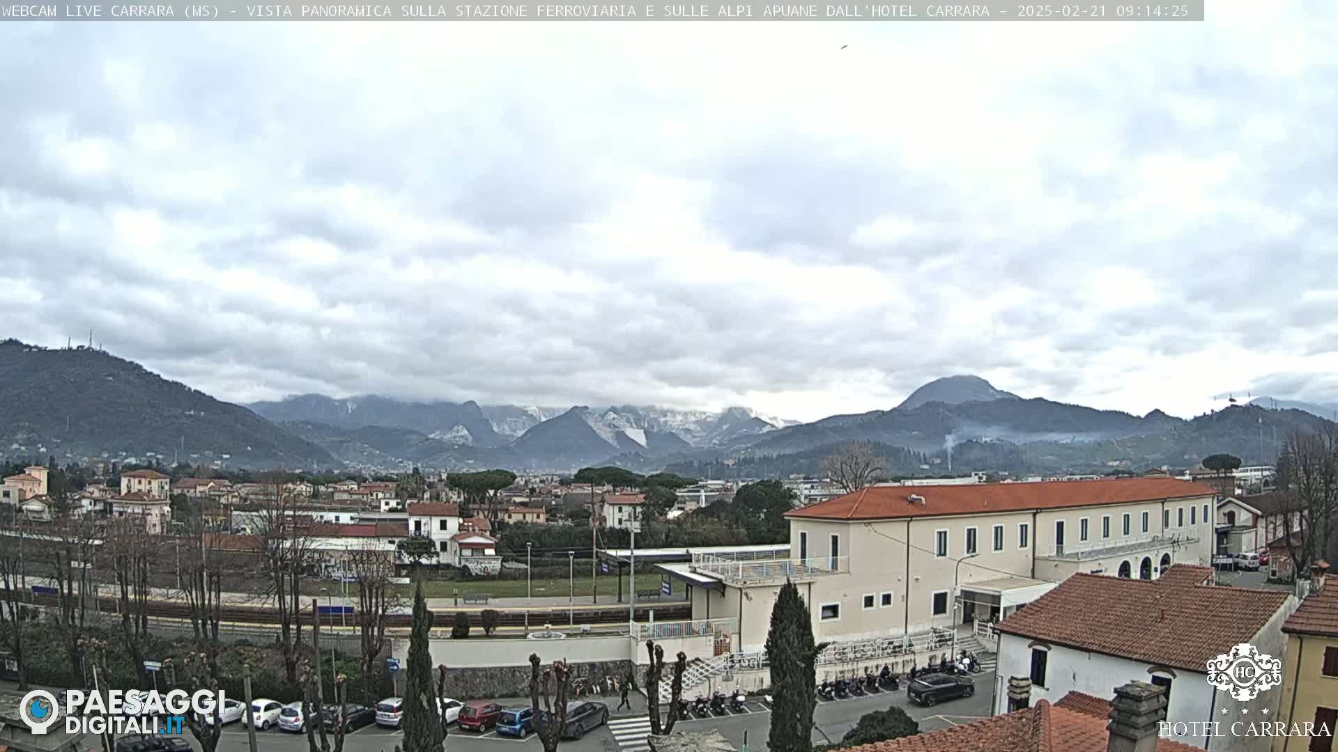A panoramic view shows a town nestled in a valley between snow-capped mountains under a cloudy sky.