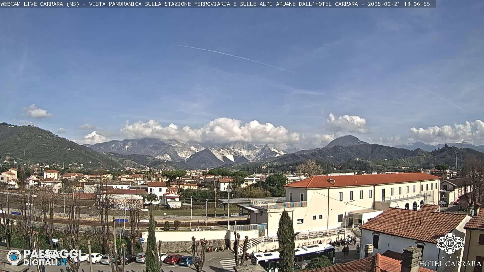 A partly cloudy day reveals a panoramic view of a town nestled beside a mountain range, with a train station and buildings in the foreground.