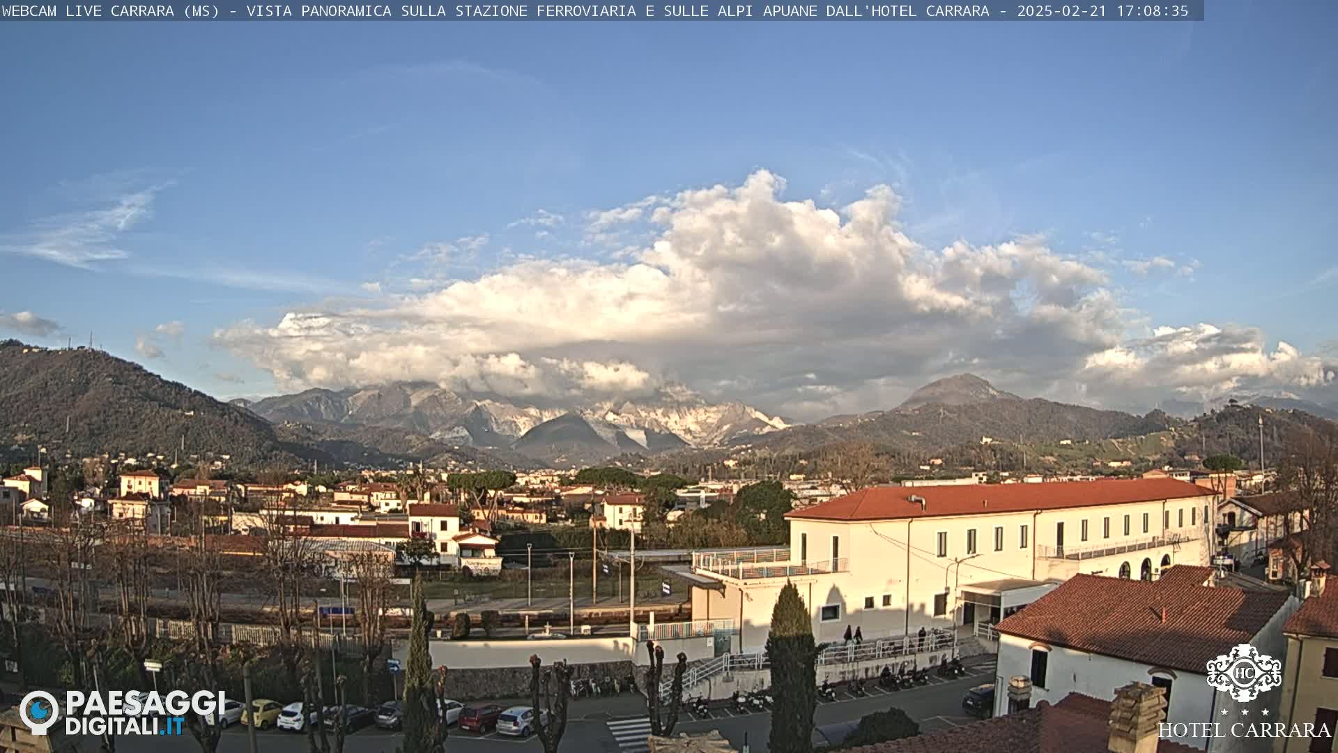 A partly cloudy day reveals a view of a town nestled in a valley, with snow-capped mountains in the background.