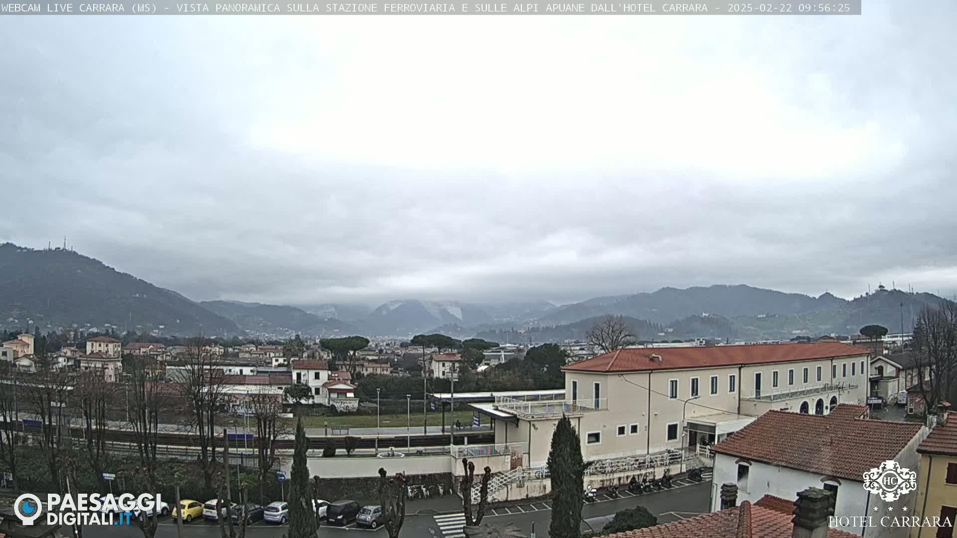 A cloudy day reveals a cityscape nestled among mountains, with a train station and parking lot visible in the foreground.