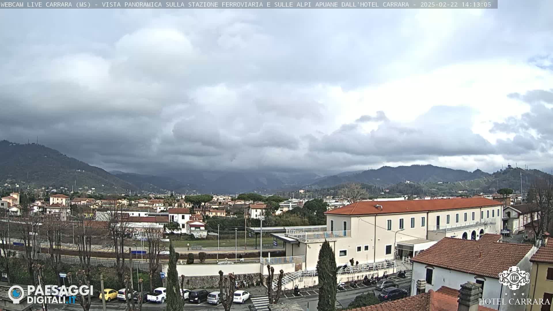 A panoramic view of a town nestled between mountains under a cloudy sky shows a train station and several buildings, including a large hotel.