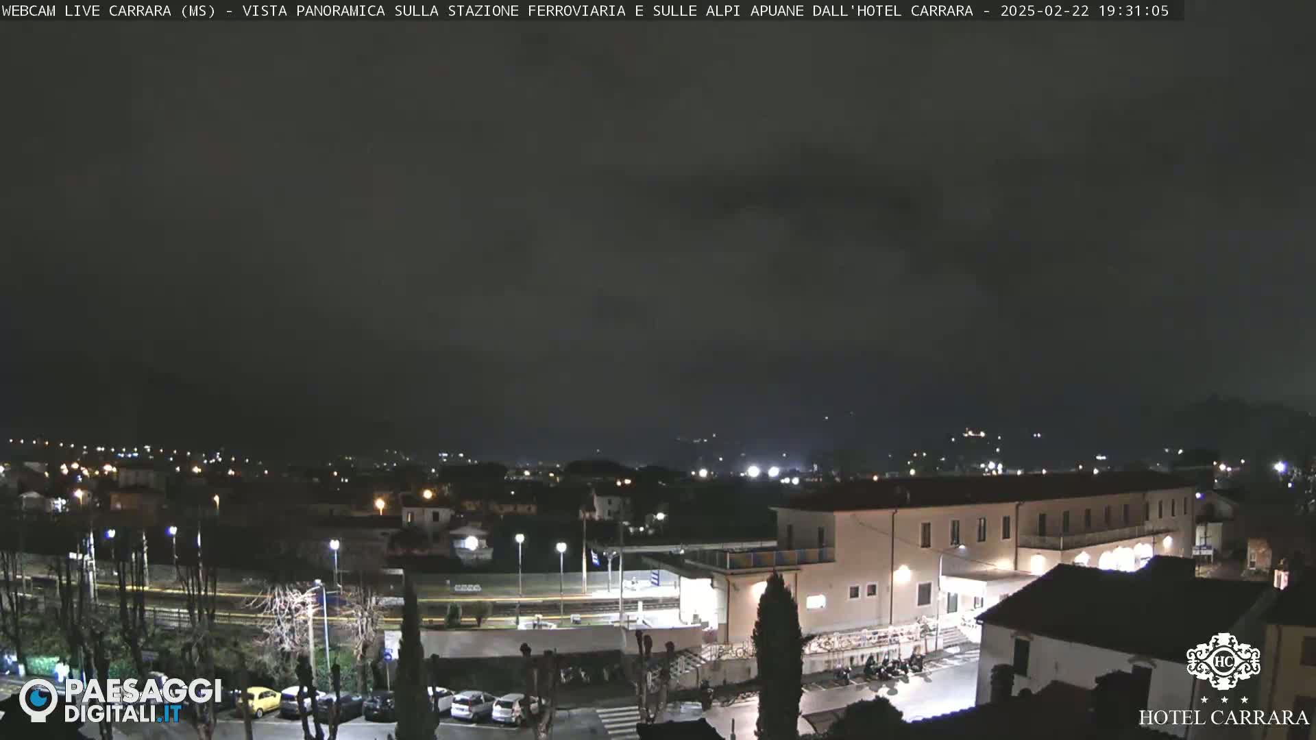 A nighttime, overcast view of a town's outskirts, featuring a train station and a hotel, with some parked cars visible.