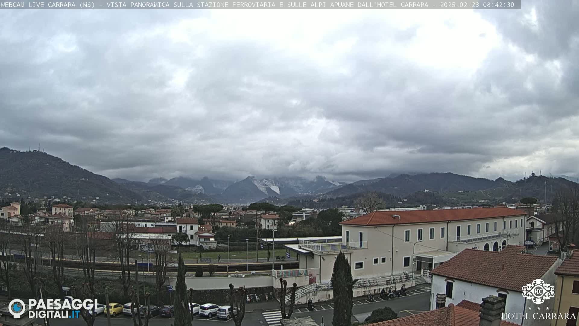 A cloudy day reveals a view of a town nestled between mountains, some with snow-capped peaks, near a train station and buildings.