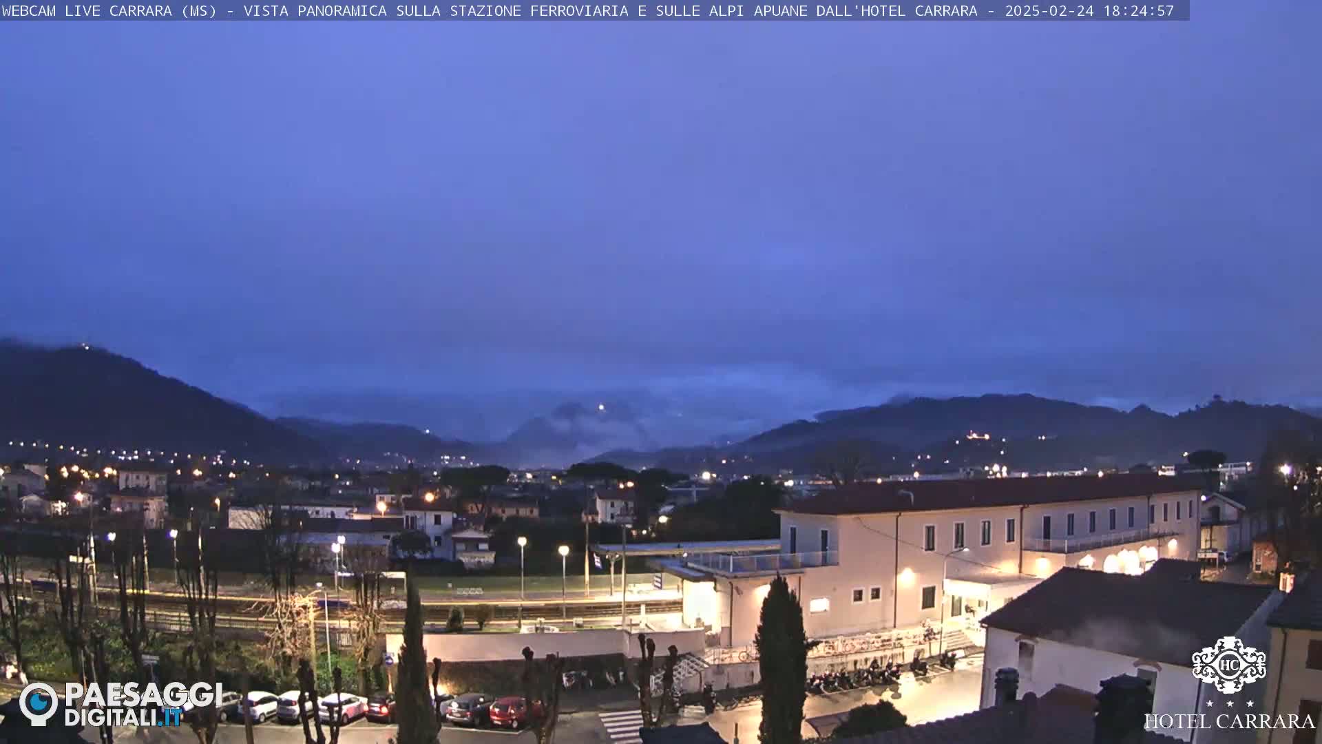 A twilight view of a town nestled in a valley between mountains, under a mostly clear, dark-blue sky.