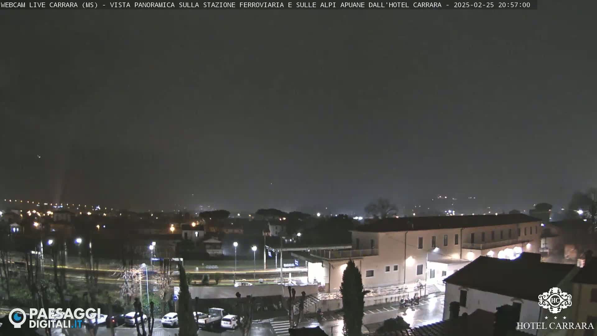 A nighttime view of a town with a large building, possibly a hotel, under a dark, slightly hazy sky with light rain.