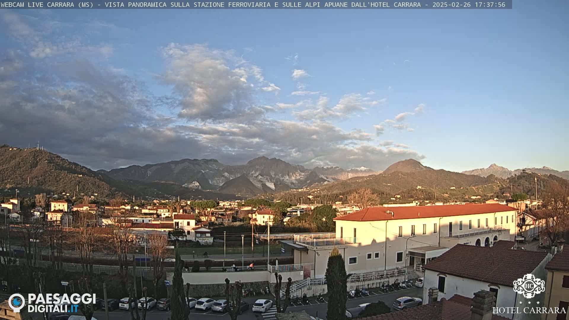 A partly cloudy daytime view shows a town nestled in a valley, with mountains in the background.