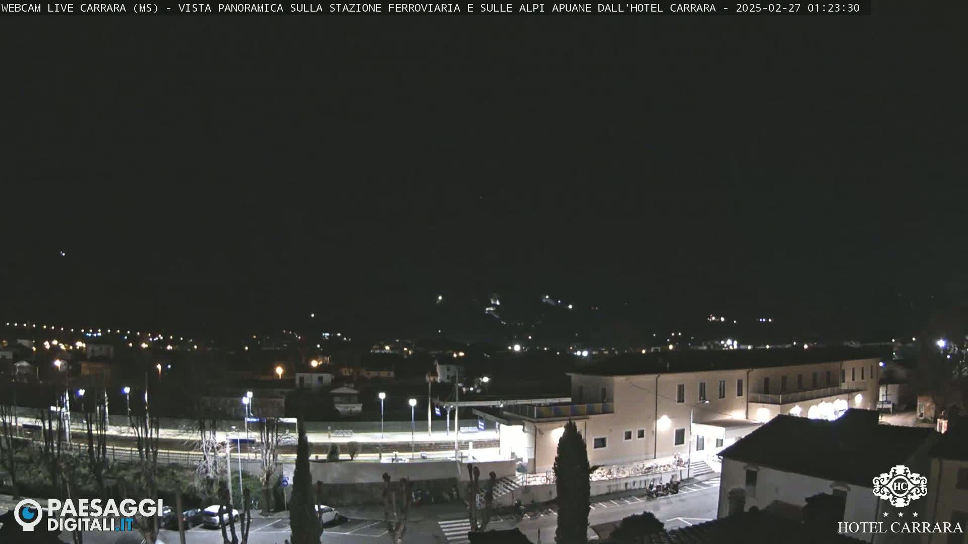 A nighttime, panoramic view of a town's train station and surrounding buildings under a clear, dark sky.