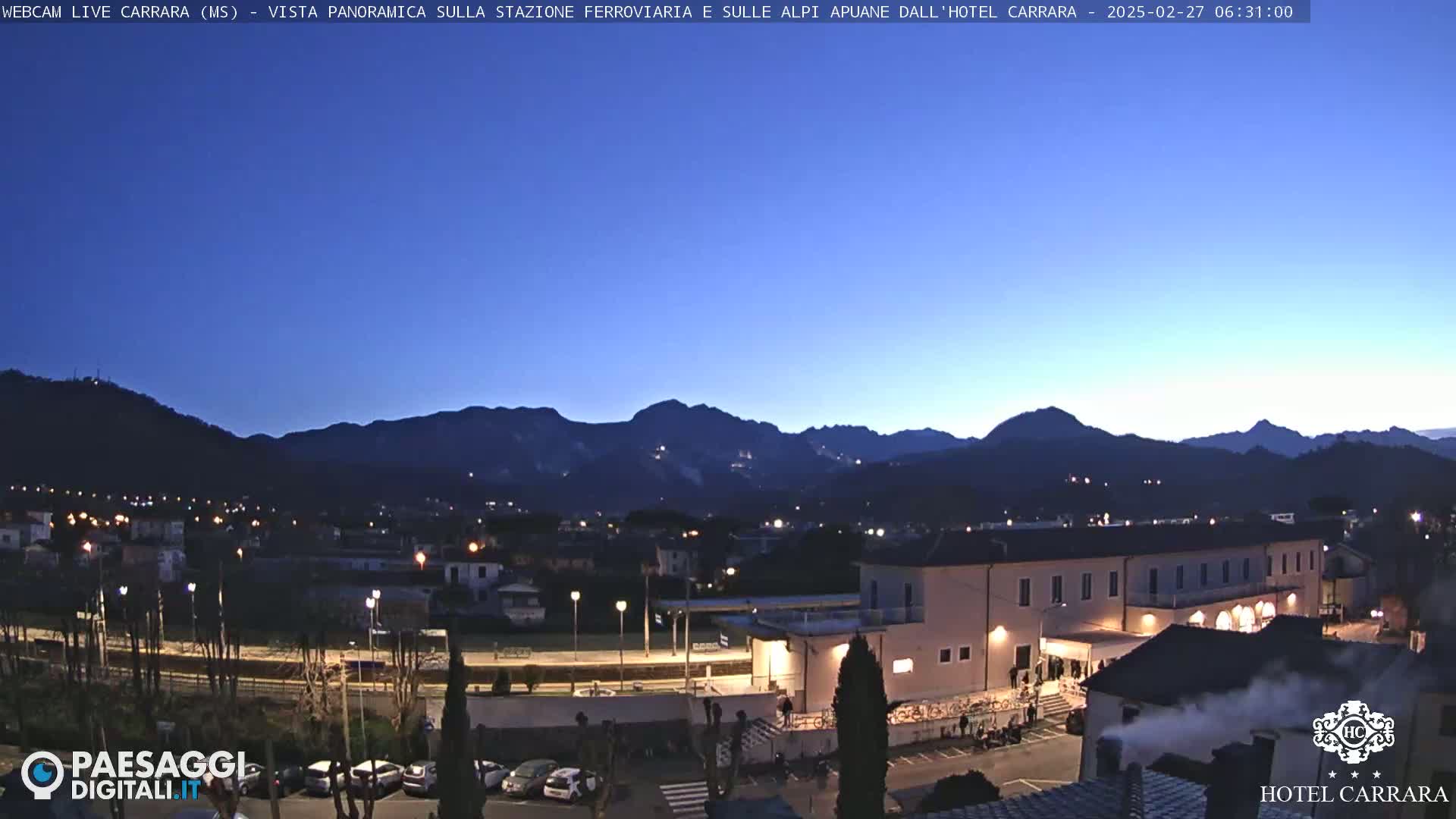 A twilight view of a town nestled in a valley, with mountains in the background under a clear, dark blue sky.