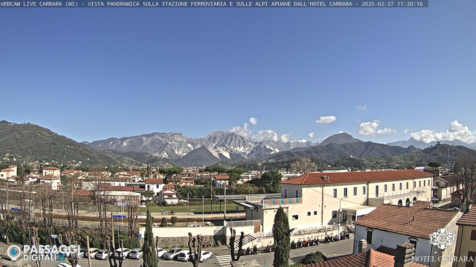 A clear day reveals a town nestled among rolling hills, with a prominent mountain range in the background and a train station in the foreground.