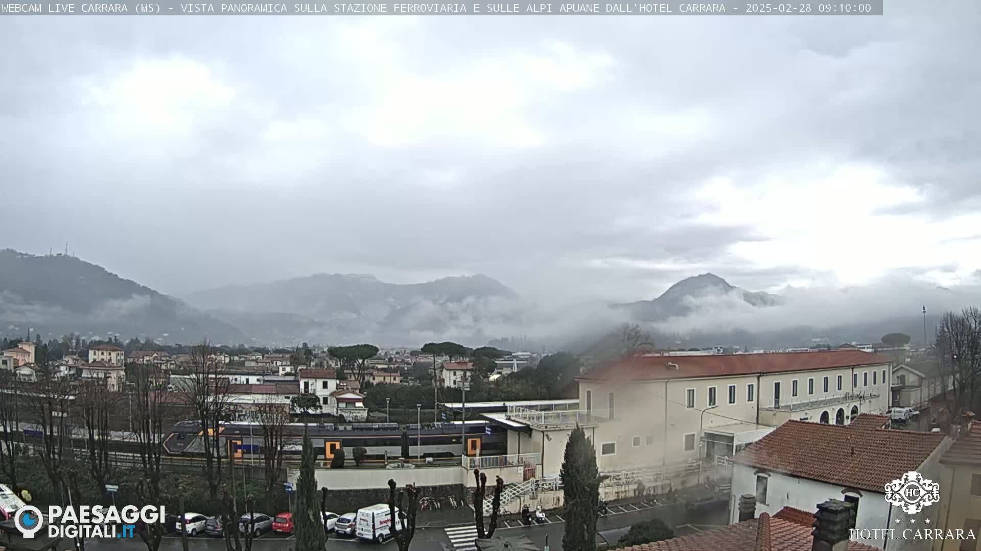 A train sits in a station near a town nestled at the foot of misty mountains under an overcast sky.