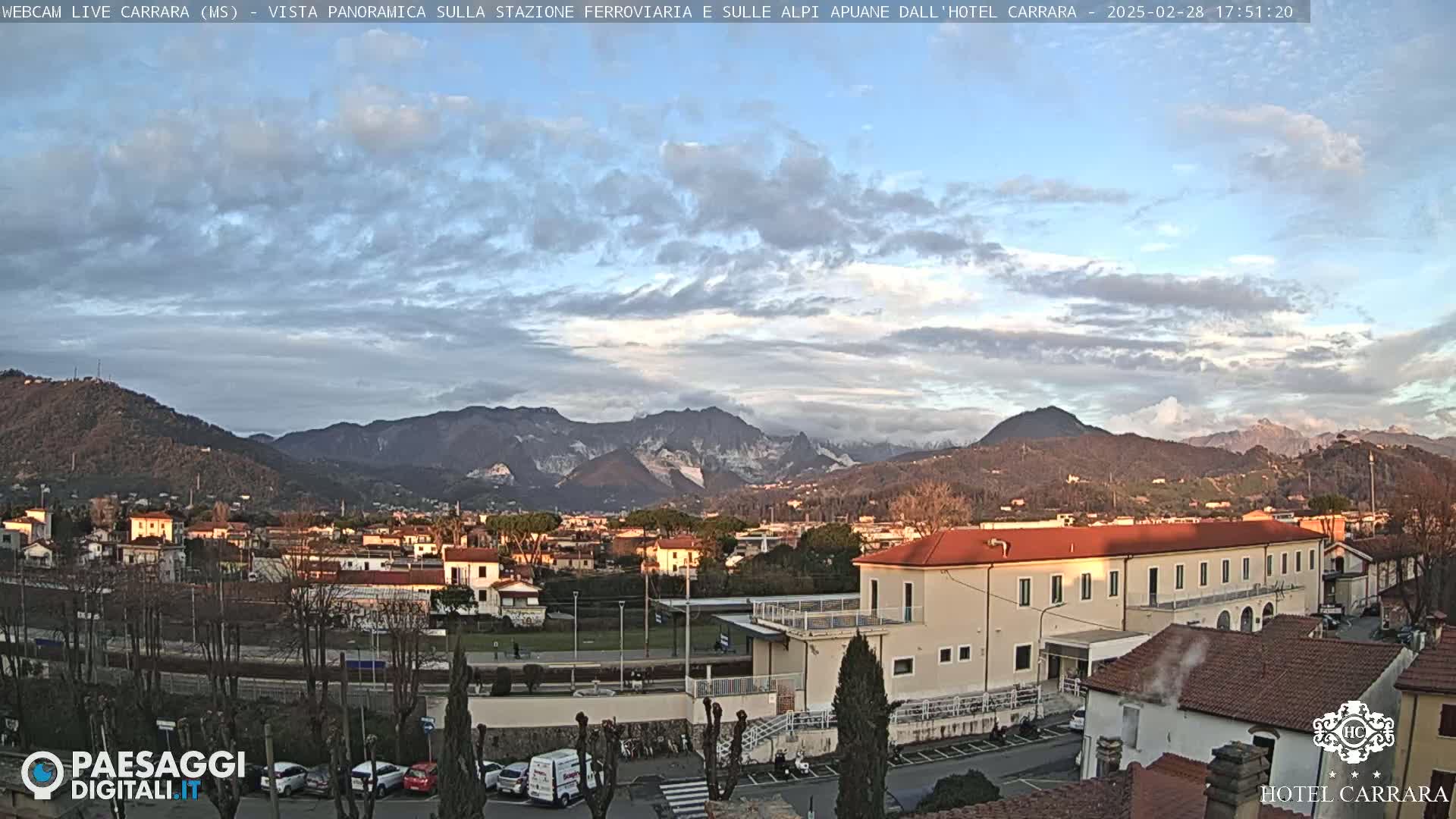 A partly cloudy sunset view shows a town nestled in a valley, with snow-capped mountains in the background.