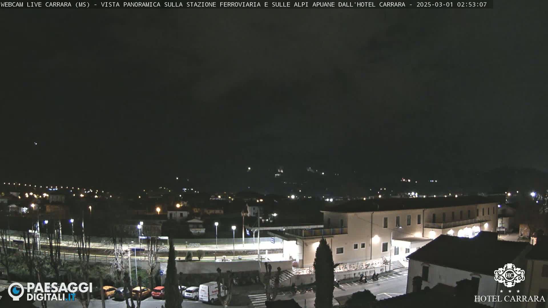 A nighttime, panoramic view of a town with a train station and a large hotel, under a dark, cloudy sky.