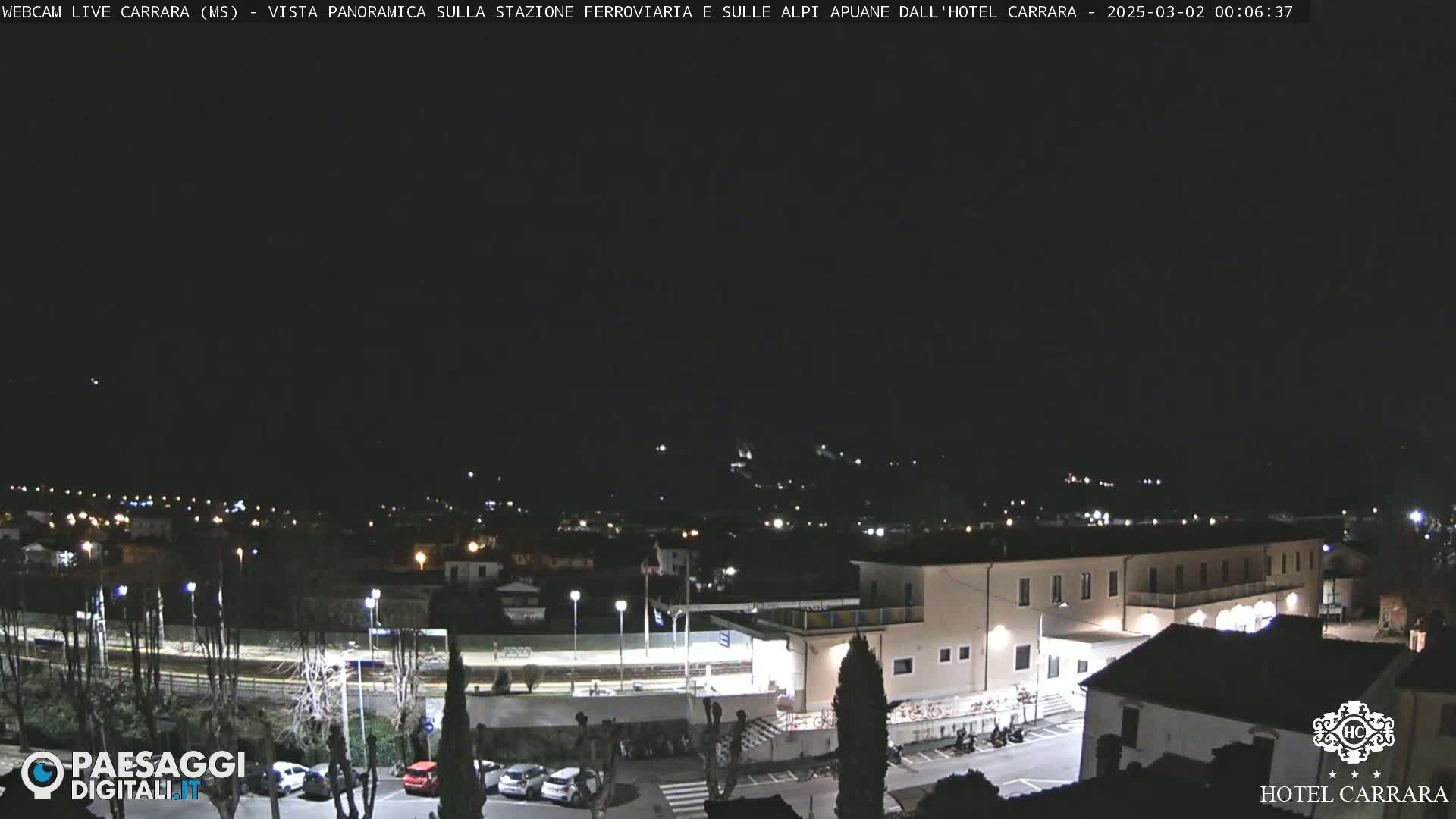 A nighttime, panoramic view of a town, showing a train station, buildings, and a parking lot under a clear, dark sky.