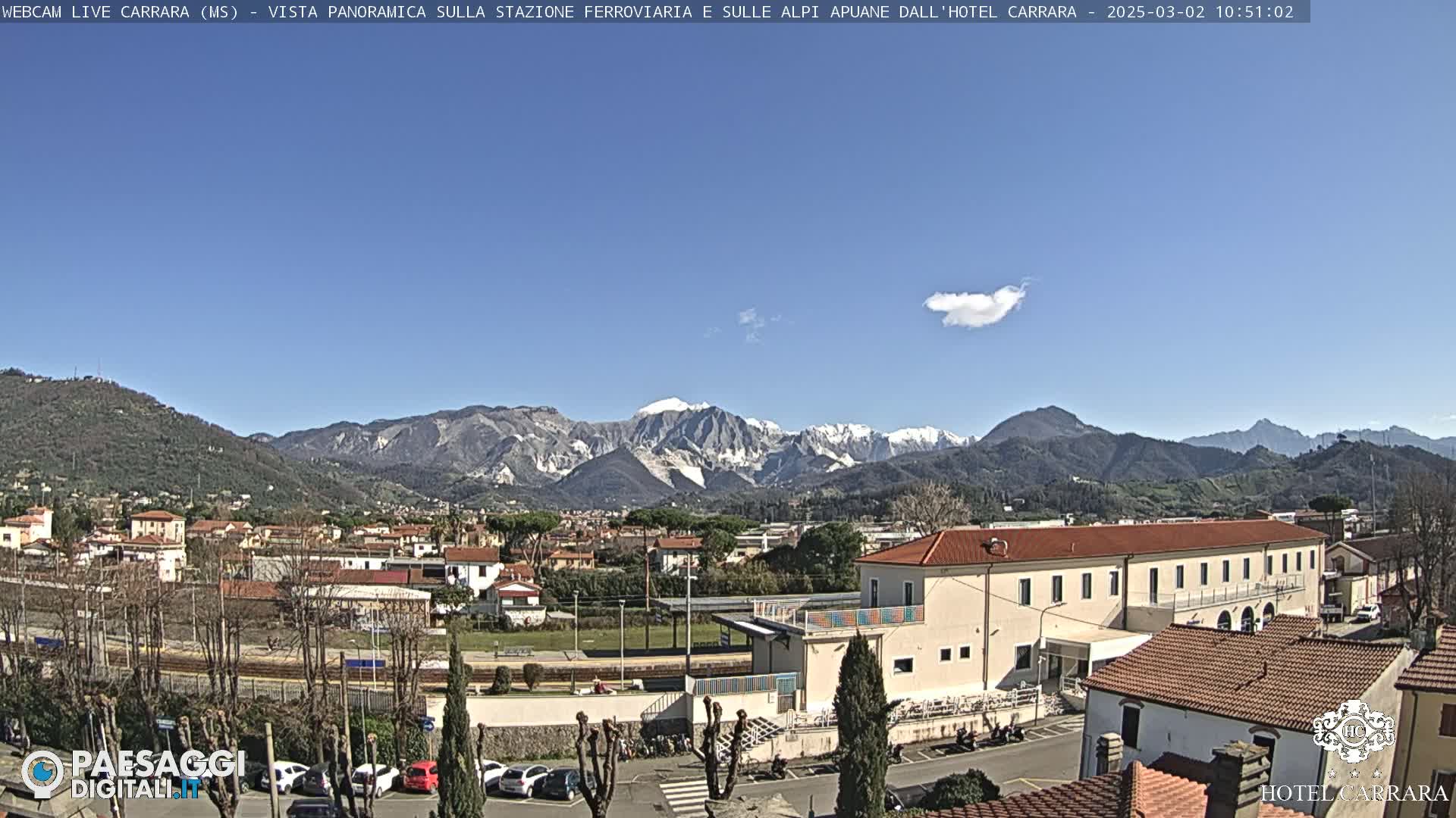 A clear, sunny day reveals a snow-capped mountain range in the distance, overlooking a town with a train station and various buildings, including a large hotel.