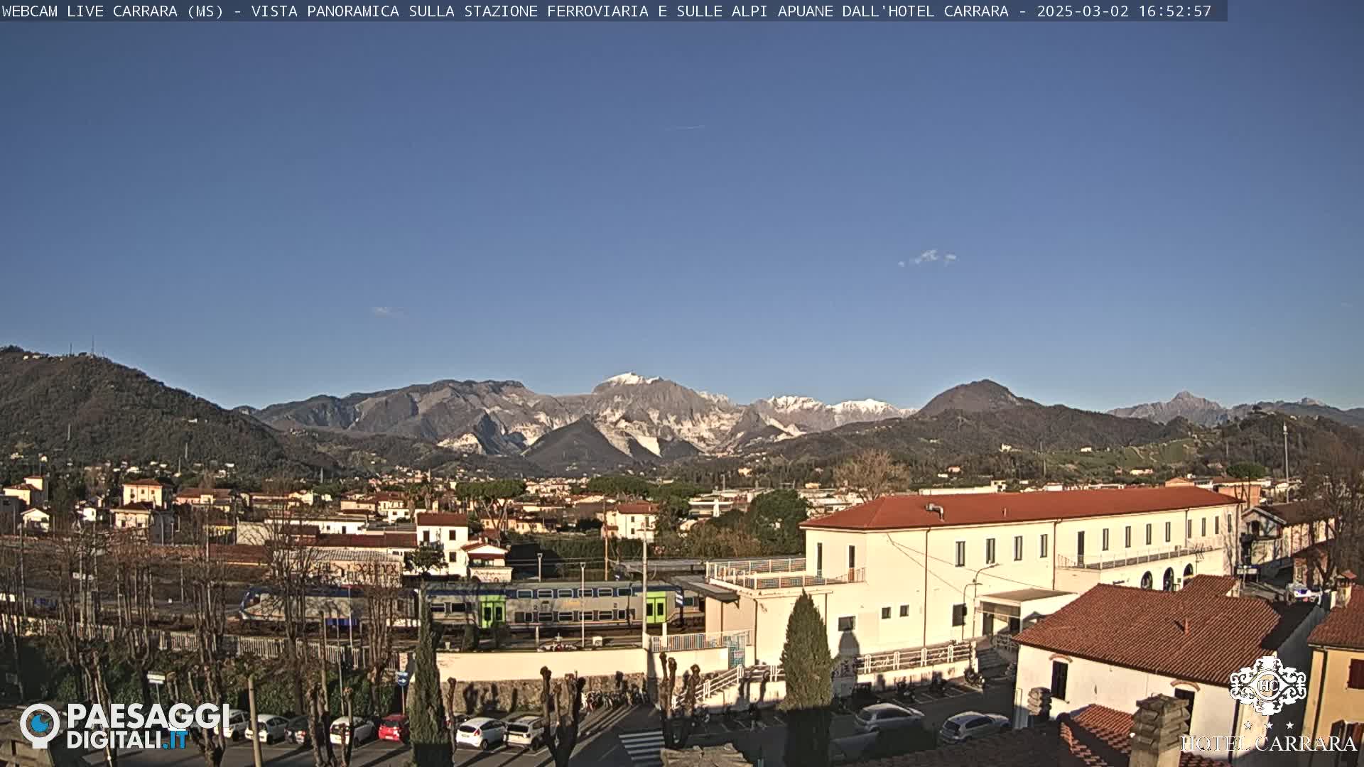 A long, low building sits in a town nestled at the base of snow-capped mountains under a clear blue sky.